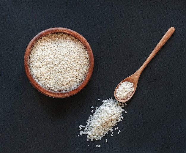 White round rice in a wooden bowl with a wooden or bamboo spoon on black background, top view