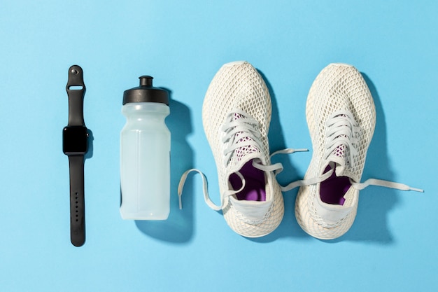 White running shoes, a sports watch and a water bottle on a blue background under the natural light of the morning sun. Concept of jogging, running, hard workout. Banner. Flat lay, top veiw