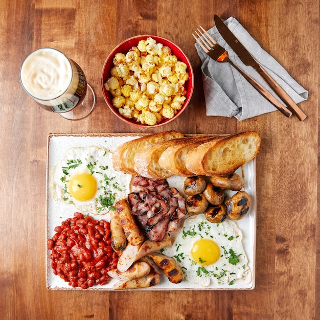 White set tray with food, sausages, bacon, beans, meatballs, scrambled eggs with greens, kvass, white toasted bread, beer in a glass goblet, bowl of popcorn