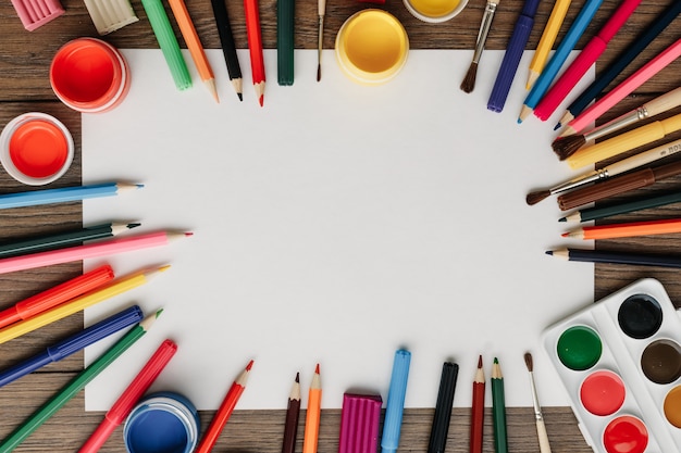 A white sheet of paper lies on a wooden brown table frame next to paints, brushes, colored pencils