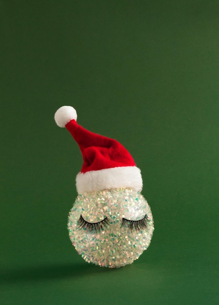 White shiny Christmas bauble with eyelashes and Santa Claus hat on bold green background Happy New Year background