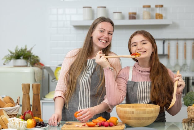 In a white spotless modern kitchen a smiling mother and daughter demonstrate how to prepare veggies for dinner