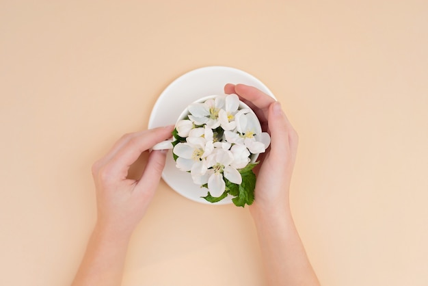 White spring apple trees blooming flowers in a coffee cup in female elegant hands on a beige background. Spring summer concept. Flat lay.