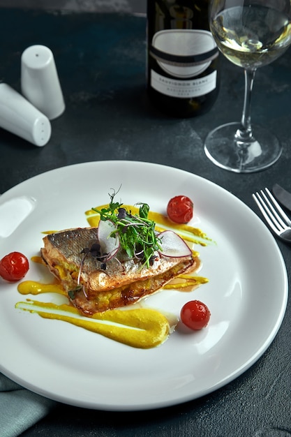 White steak of grilled fish on pillow of vegetables and yellow sauce, with tomatoes and fresh herbs. Fish on white plate with white wine on dark beautiful background. Close-up. Space