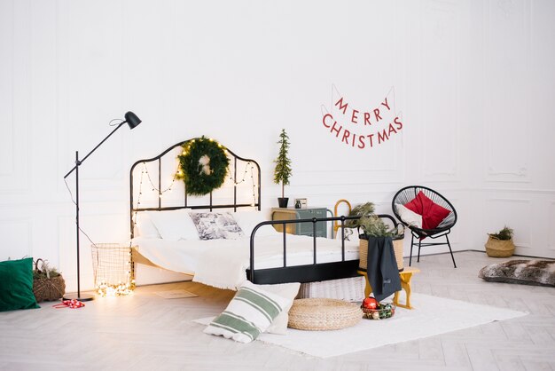 White stylish cozy scandinavian bedroom. Christmas holiday decorations: little christmas tree, wreath, led garland lights.