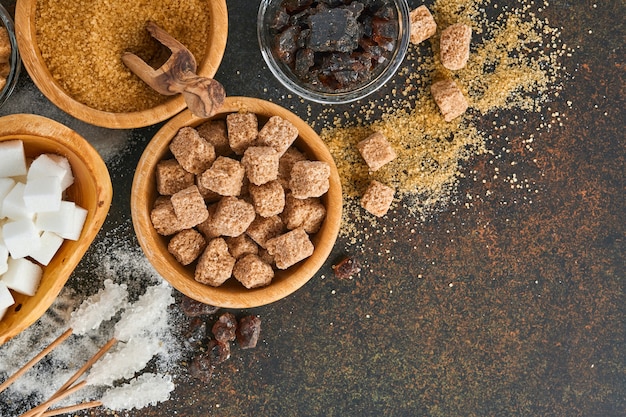 White sugar, cane sugar cubes, caramel in bamboo bowl on dark brown table concrete background. Assorted different types of sugar. Top view or flat lay.