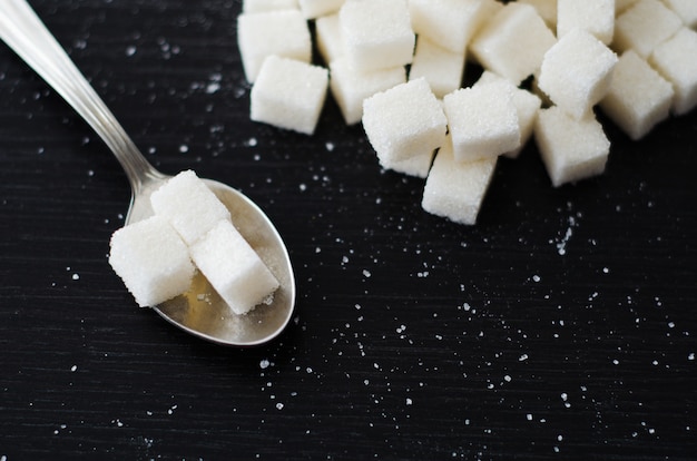 Foto cubetti di zucchero bianco ammucchiati nel mucchio sulla scrivania nera con cucchiaio