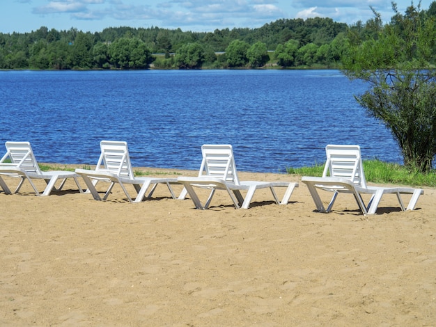 Foto sedie a sdraio bianche sulla riva sabbiosa delle vacanze estive del bacino idrico. copia spazio.