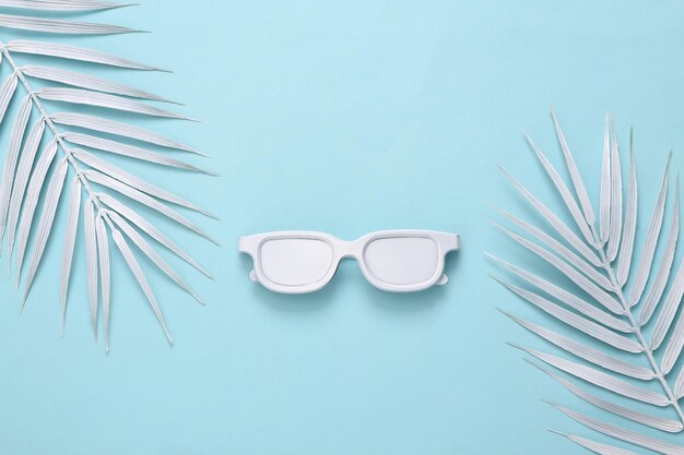 Белые солнцезащитные очки и пальмовые листья на синем фоне Минимальное путешествие в тропическом стиле Вид сверху