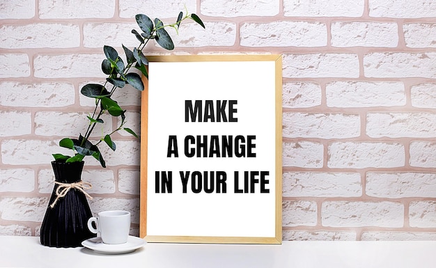 On a white table against a light brick wall a branch of eucalyptus in a dark vase a white cup and a light wooden frame with the text MAKE A CHANGE IN YOUR LIFE Home office interior