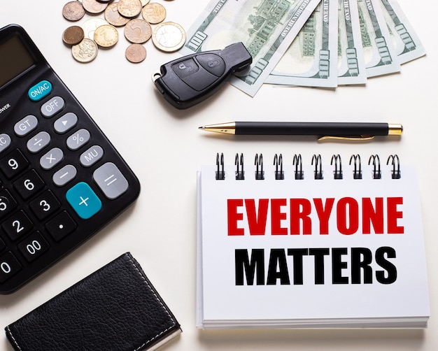 On a white table is a calculator, car key, cash, a pen and a notebook with the inscription EVERYONE MATTERS