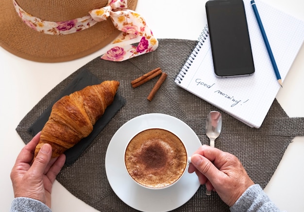 A white table with a sweet breakfast. Homemade cappuccino with cinnamon powder and a fresh croissant. Smartphone - old  lifestyle and new technology