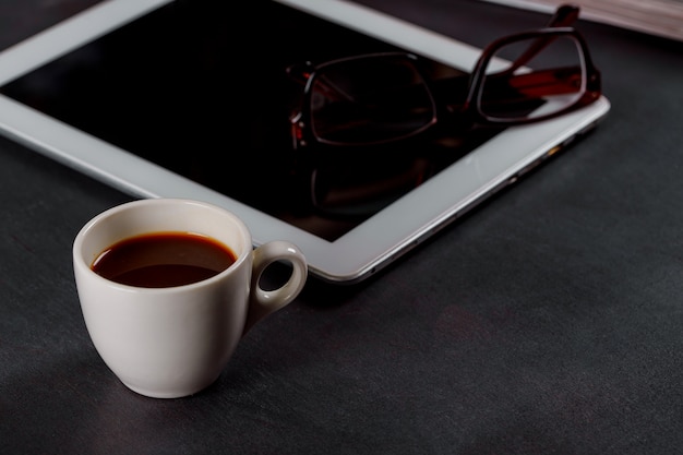 White tablet computer on a wooden table with a glasses, a cup of black coffee