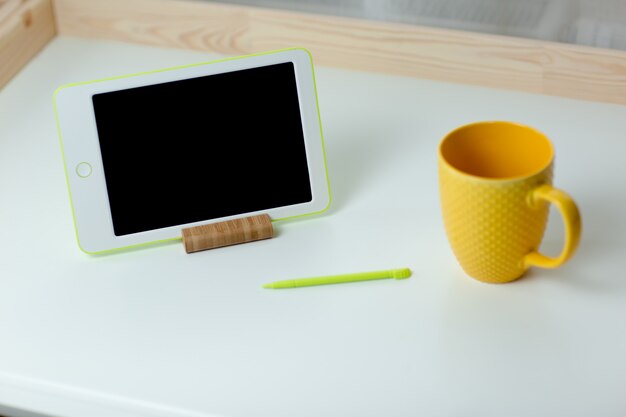 White tablet and yellow cup of tea on white table. Home office while self-isolation, working from home. Online education, e-learning while quarantine.