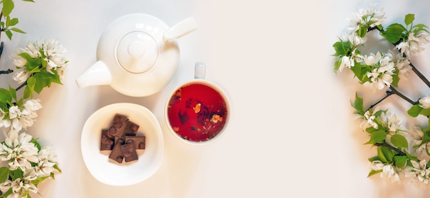 White tea pot, red fruit tea, chocolate, apple tree blossom, white wall flat lay. Floral spring flowers concept