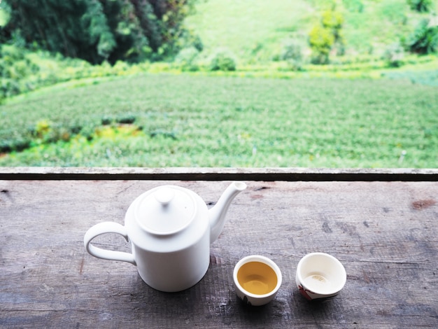 White teapot and cups on vintage wooden table over green tea farm .