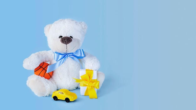 White teddy bear holding a  tiny ball small present and toy car on light blue background