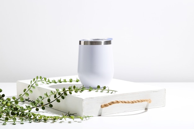 Белая термочашка с зеленым растением на деревянном подносе, минимальная композиция для презентации дизайна