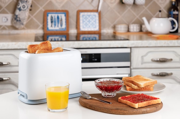 White toaster glass of orange juice slices of toasted bread crispy toast with raspberry jam and bowl with confiture on wooden board Served table for delicious meal Fresh breakfast concept