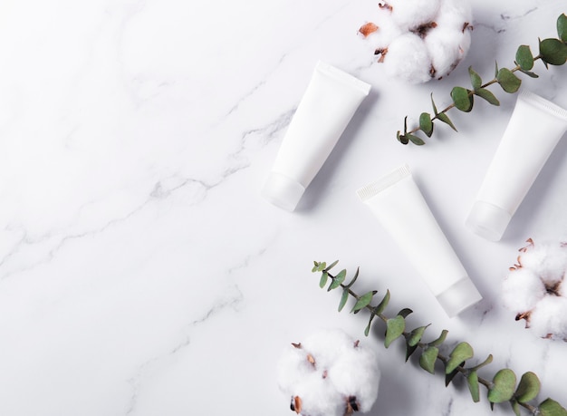 White tubes of cream on a marble wall with eucalyptus and cotton flowers