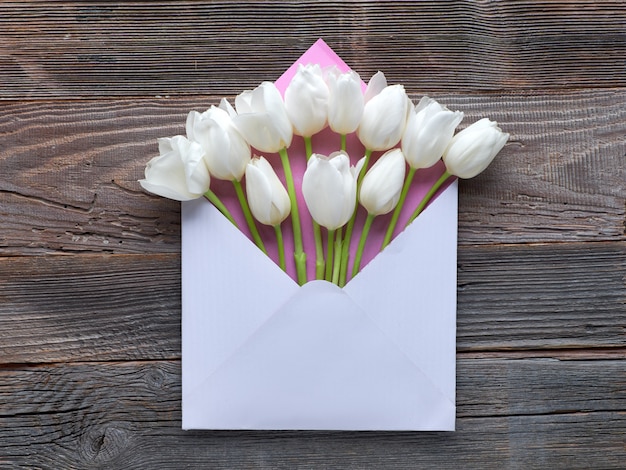 White tulips in blank paper envelope, flat lay on rustic wood