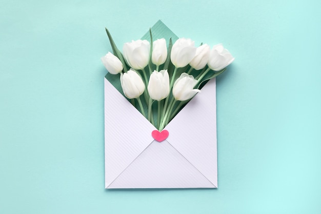 White tulips in white-green envelope on blue mint paper background, springtime flat lay