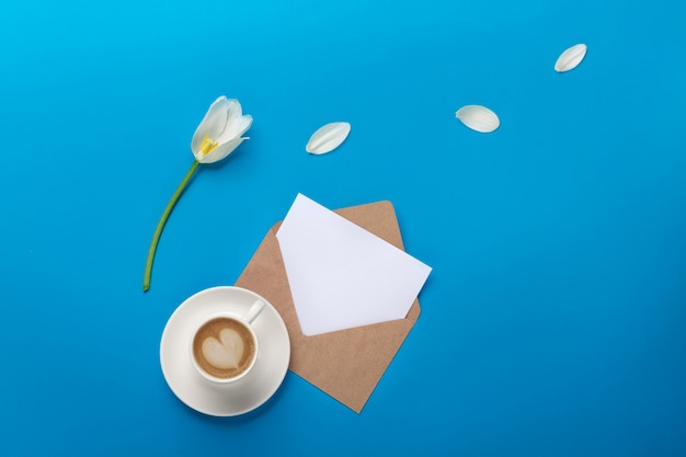 White tulips with petals, cup of coffee, a love note and envelope on a blue background