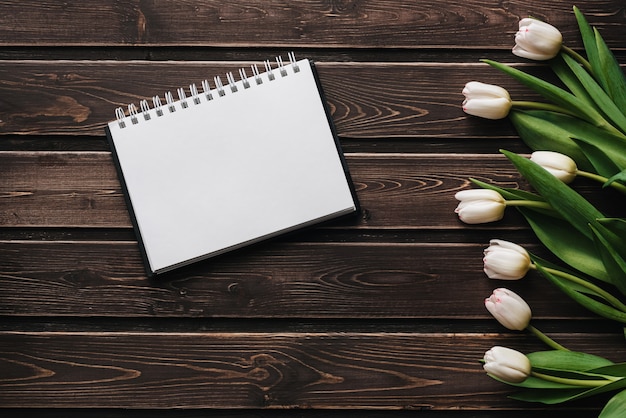 White tulips on a wooden table with an empty notebook. Flat lay, top view composition