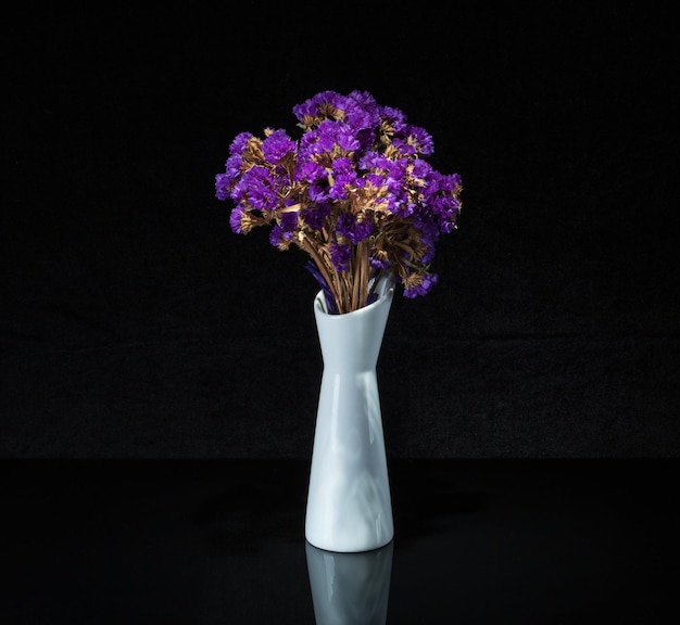 A white vase with immortelle flowers on a black background is reflected in a mirror stand
