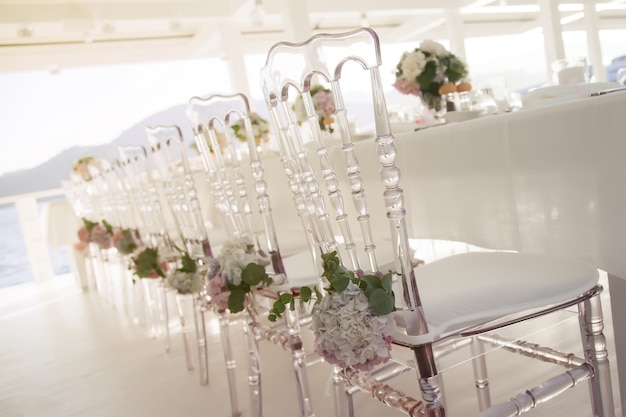 White wedding chairs decorated with fresh flowers in restaurant with panoramic view of sea. Luxury design for event. Floral chair decorations for outdoor parties and dinners. Copyright space for site