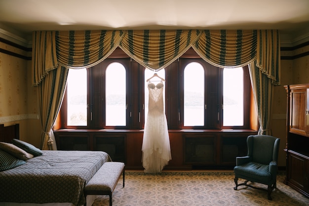 White wedding dress is hanging by the window in a hotel room