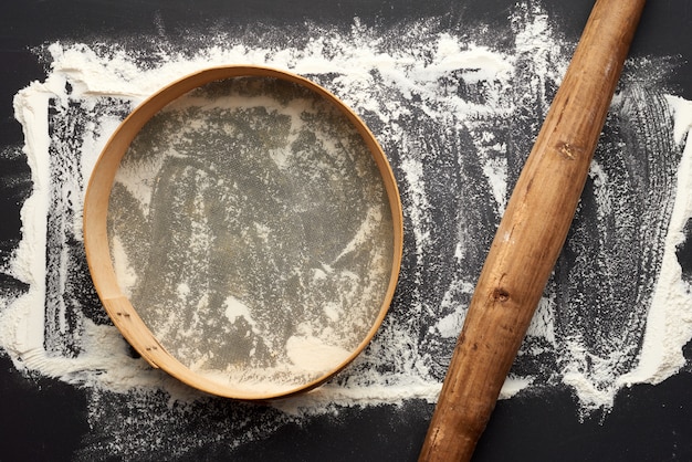 Photo white wheat flour scattered on a black table and very old brown wooden rolling pin