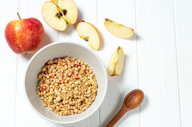 White wheat granola in a white bowl in a composition with a spoon and apple on white wooden background. 