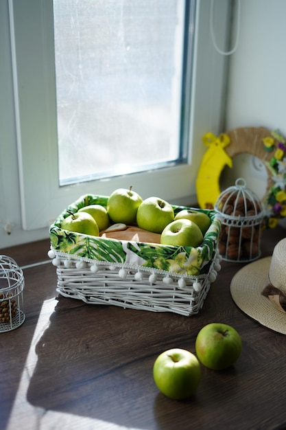 A white wicker basket with green apples on a wooden table near the window