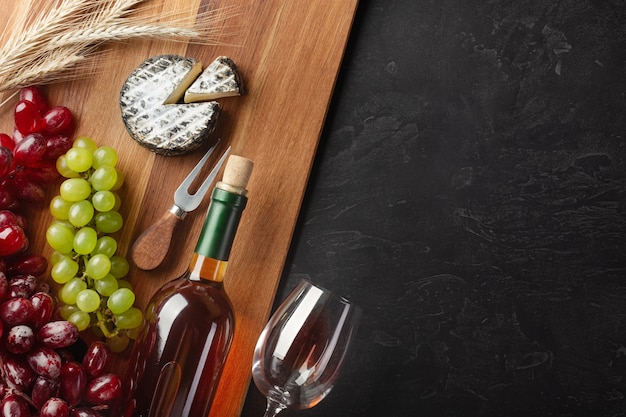 Бутылка белого вина, гроздь винограда, сыр, колосья пшеницы и рюмки на деревянной доске и черном фоне. Вид сверху с копией пространства.