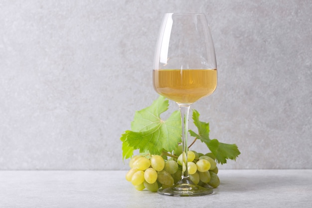 White wine in a glass and a bunch of grapes on the table. Light wall.