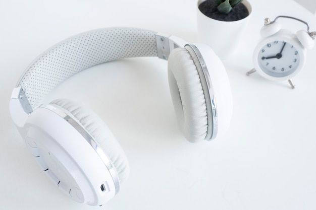 White wireless headphones and a small white alarm clock on a white table.
