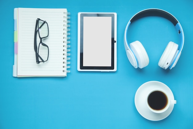 White wireless headphones, tablet, glasses, coffee on the blue background