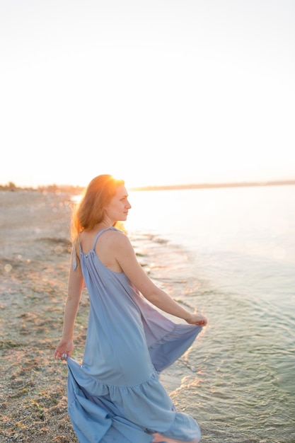 파란색 드레스를 입은 백인 여성이 새벽에 해변을 산책합니다. 바다와 석양