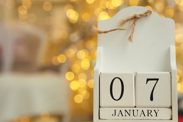 White wooden calendar with cubes and date January 07 and lights bokeh from a garland in the background.