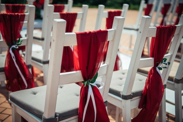 Белые деревянные стулья, украшенные красной тканью и лентами для свадебного оформления на открытом воздухе