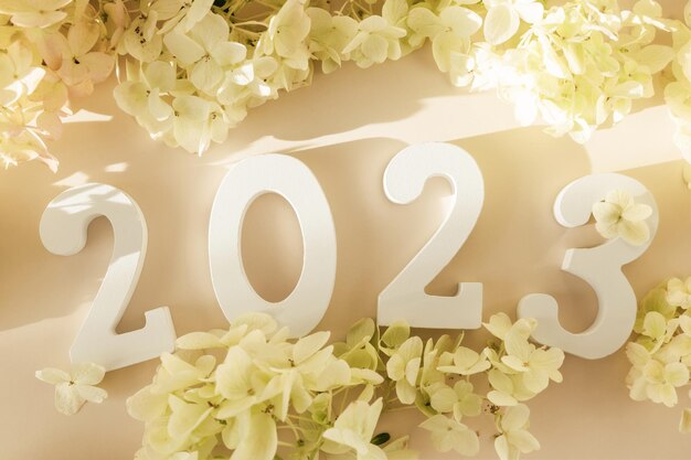 Photo white wooden number 2023 on pastel beige background with flowers top view flat lay happy new year 2023