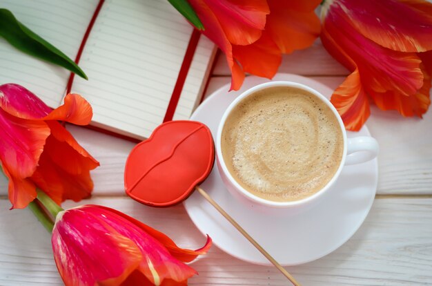 On a white wooden table is a red notebook with tulips, there is a cup of coffee and gingerbread in the form of scarlet lips