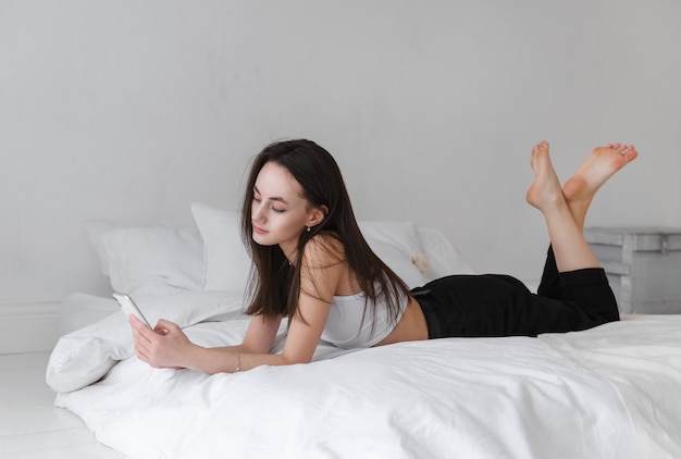 Photo white young brunette woman in white top lying on bed with phone, side view