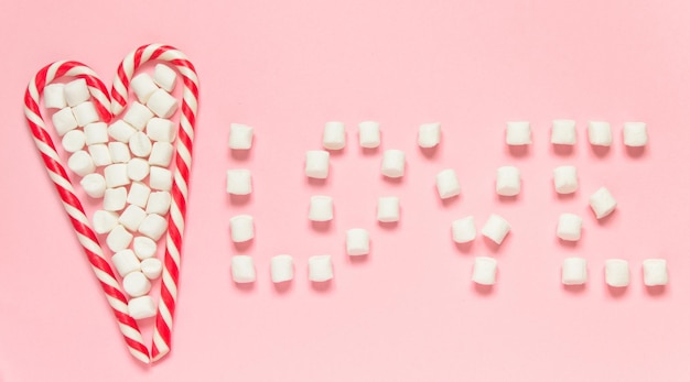 Whitered candy canes and marshmallow in heart shape and word love on pink background