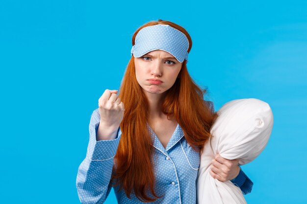 Who disturbed my sleep. Mad and displeased, angry grumpy redhead woman threaten sibling being too loud in morning, shaking fist frowning, holding pillow, wearing pyjama and sleep mask