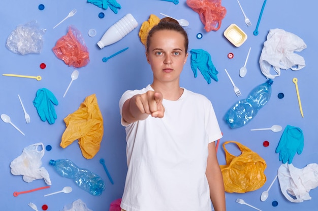 Photo who else but you portrait of dissatisfied caucasian woman activist engaged in antipollution campaign looks and pointing finger at camera posing against blue background with plastic garbage around