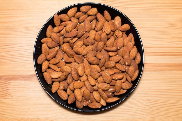 Whole almond nuts in black plate on the wooden background. Healthy vegetarian snack. Close-up photo.