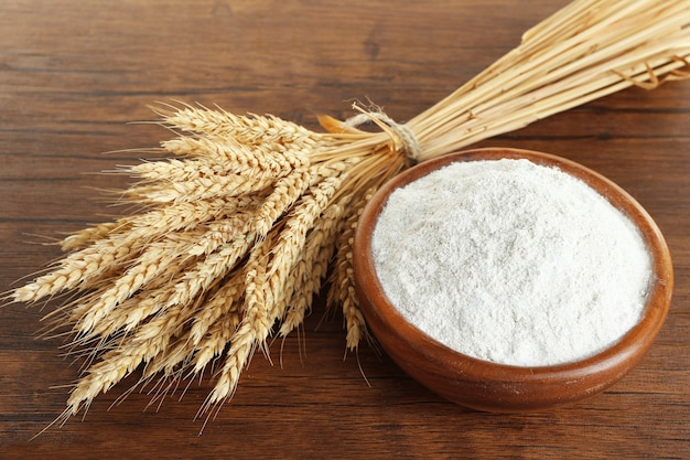 Whole flour in bowl with wheat ears on wooden table, closeup