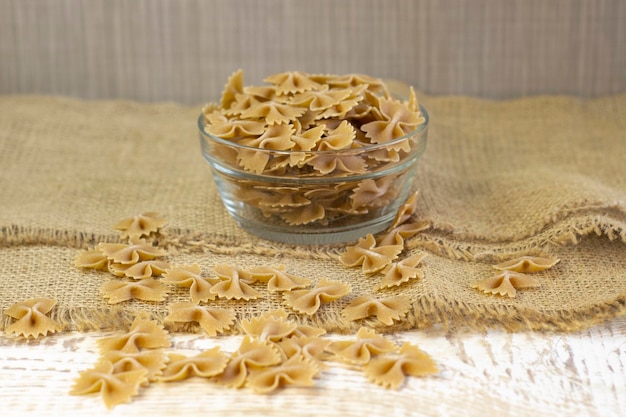 Whole grain dark pasta bows on brown plate on a rough wooden light background with selected focus